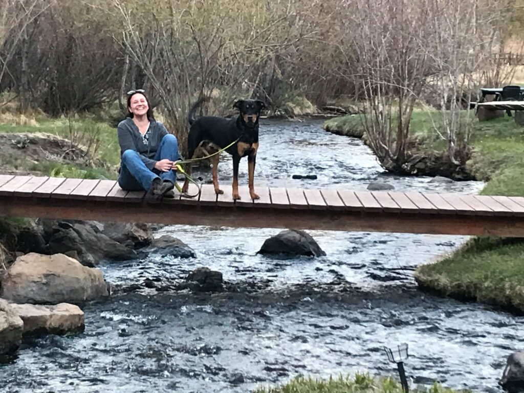 Katrina Kaye and pup - white woman and black dog sitting on a footbridge over a stream