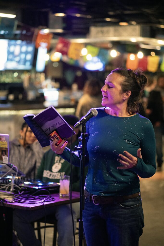 Katrina Kaye reading at an open mic - white woman in teal long-sleeve shirt at a microphone reading from a book