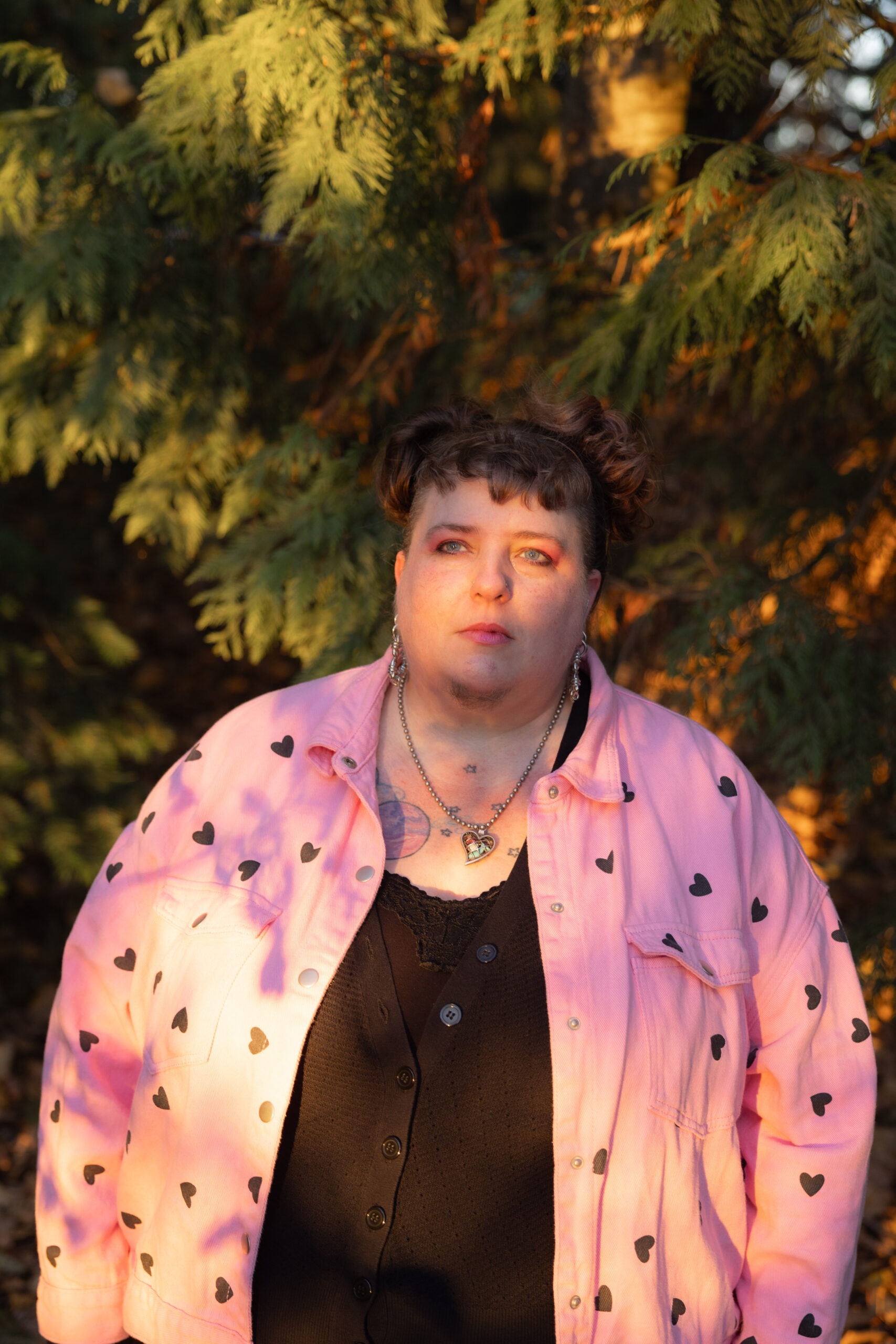 fairer-skinned queer person with brown hair pulled up, wearing a heart-shaped necklace, a pink overshirt with black hearts, and black undershirts. she/they are standing in front of fir trees.