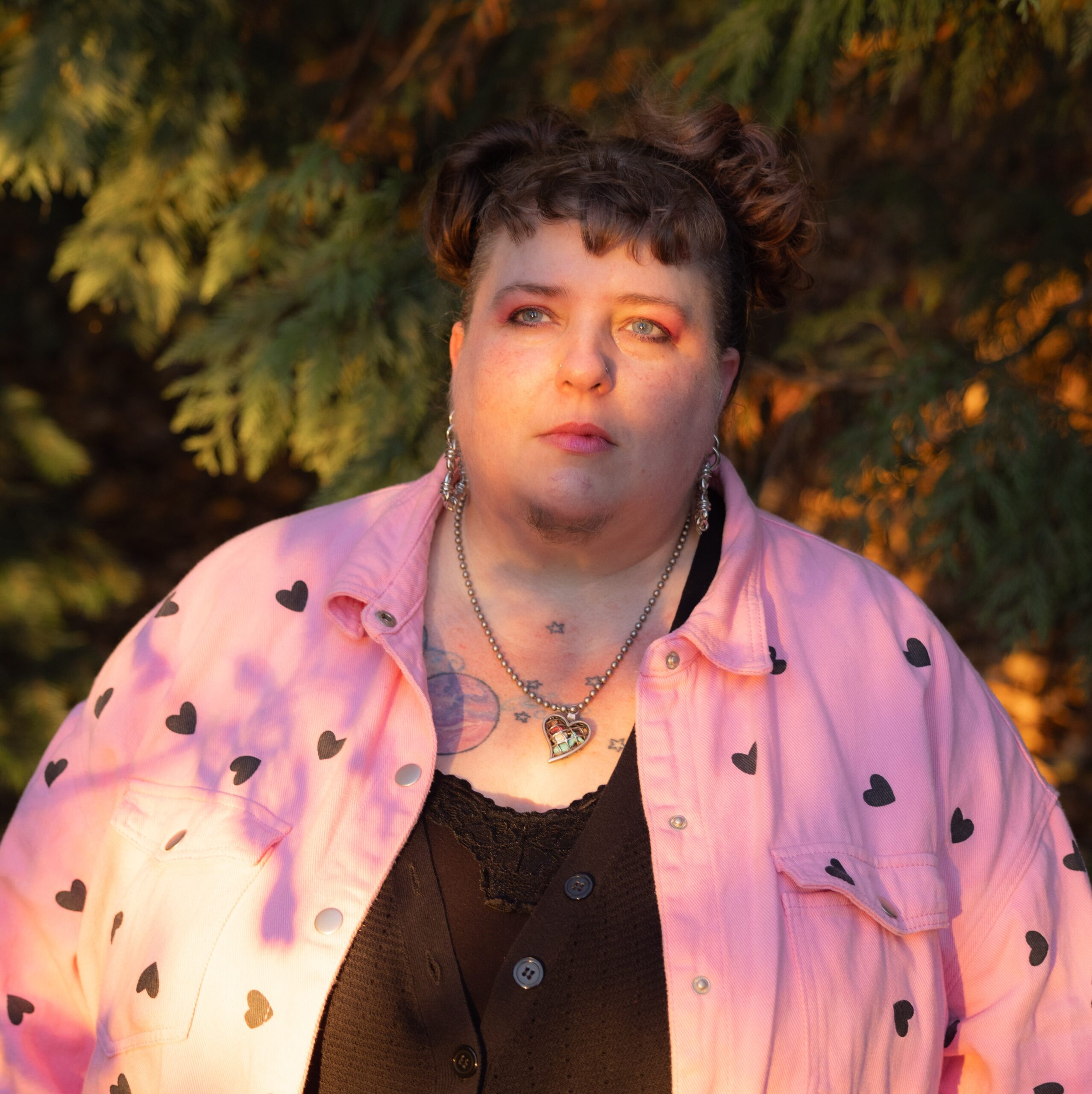 fairer-skinned queer person with brown hair pulled up, wearing a heart-shaped necklace, a pink overshirt with black hearts, and black undershirts. she/they are standing in front of fir trees.